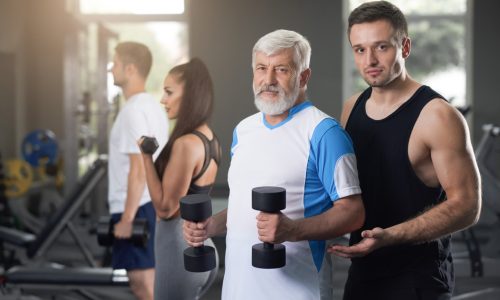 Portrait of two athletic men looking at camera. Elderly attractive fit man doing exercises in gym. Young professional coach help senior adult training with sport facilities.